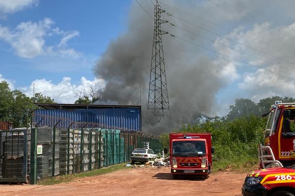 La fumée épaisse s'échappe du site de l'incendie, menaçant un pylône électrique à haute tension, tandis que les pompiers et leurs véhicules s'affairent à contenir les flammes et à sécuriser la zone.