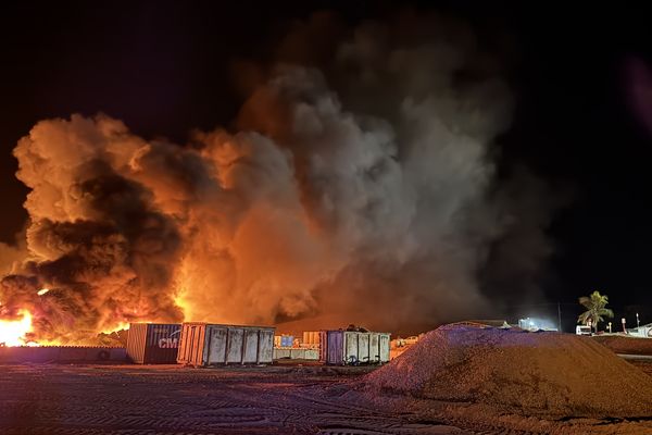 Des fumées épaisses et orange dominent le ciel alors que les pompiers tentent de contenir l’incendie, menaçant de se propager dans la zone de stockage.